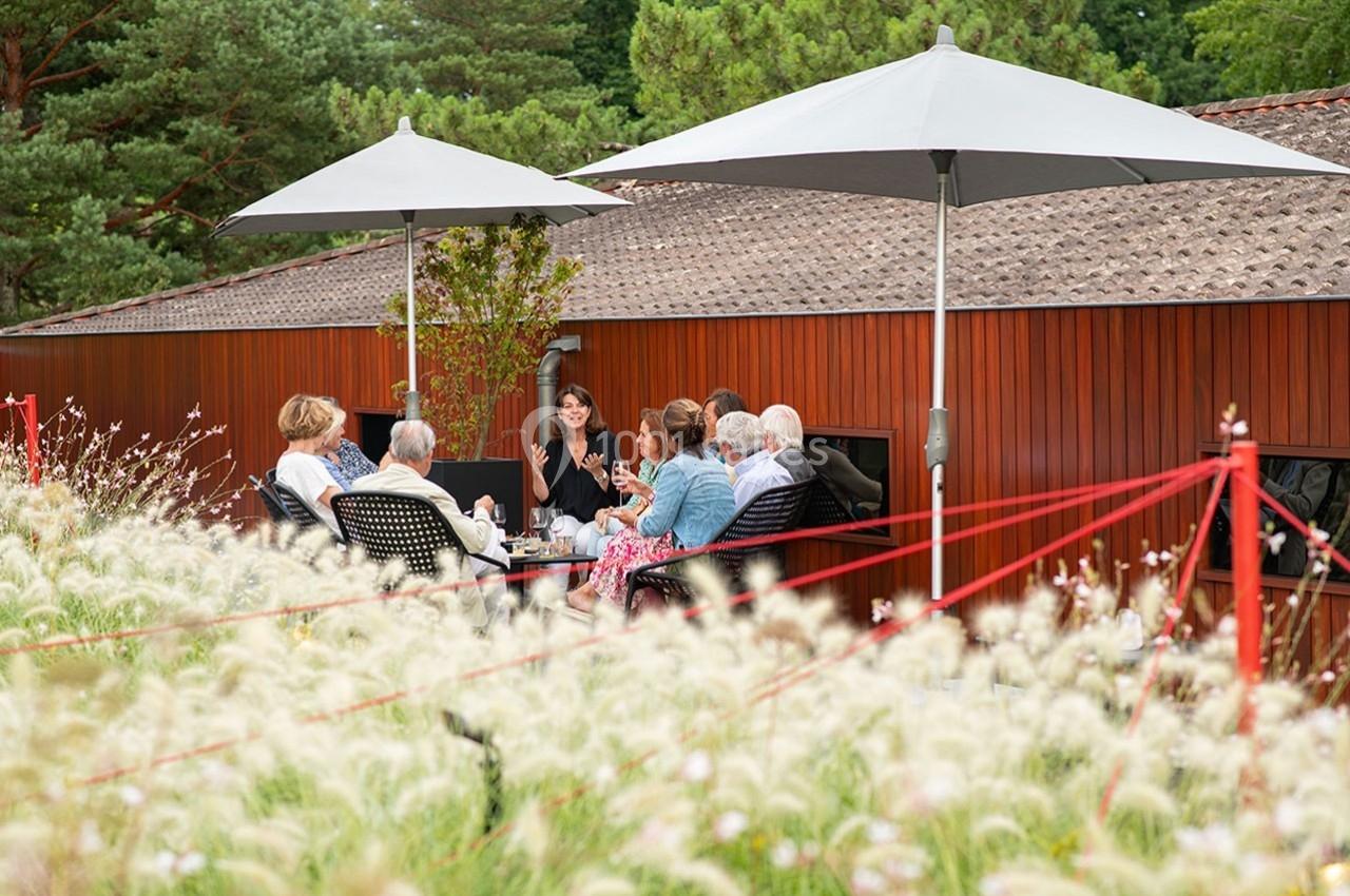 Groupe de personnes assises autour d'une table en terrasse, sous des parasols, entourées de végétation.