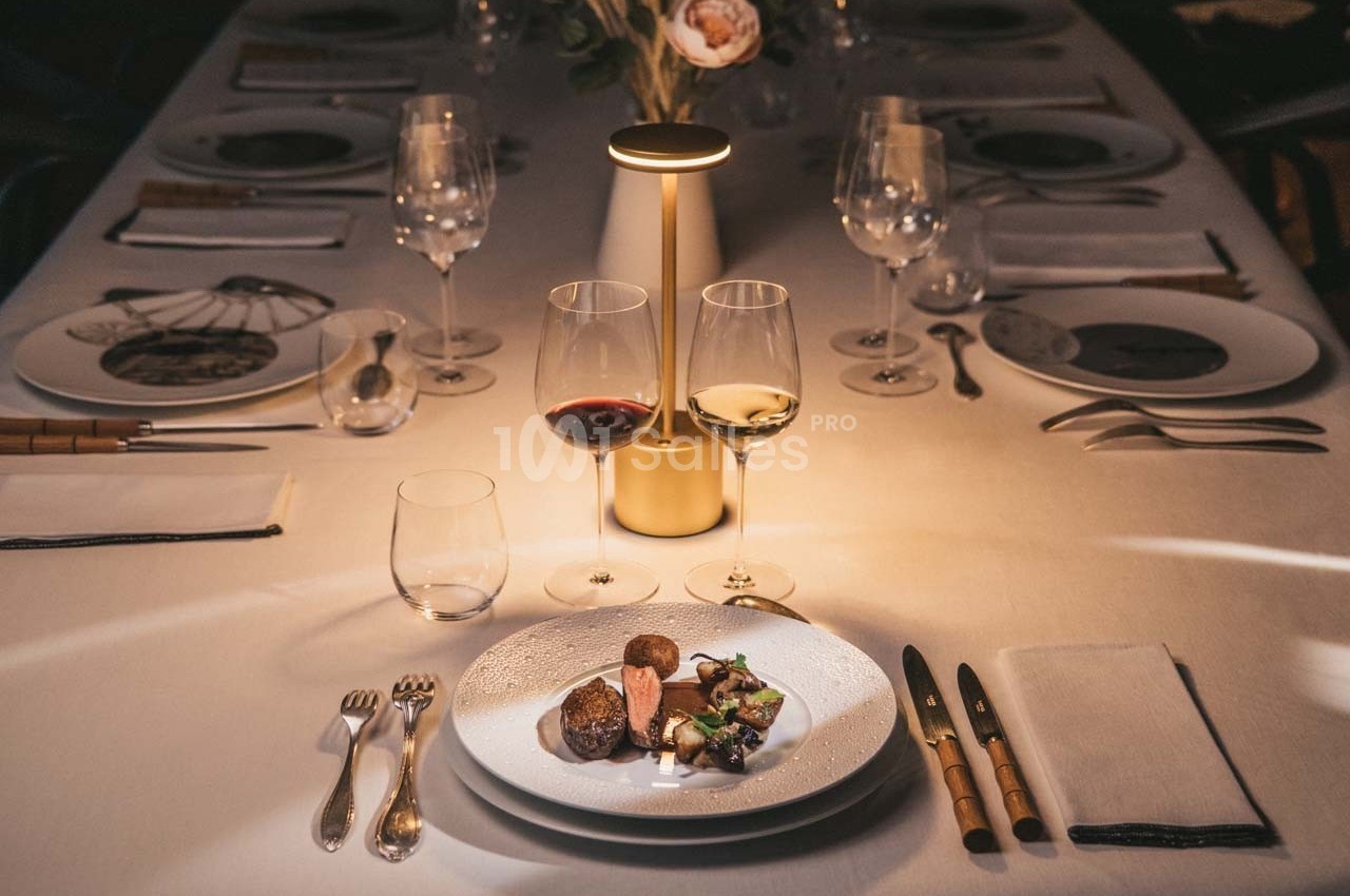 Table élégamment dressée avec assiettes, verres à vin et plat raffiné sous une lumière tamisée.