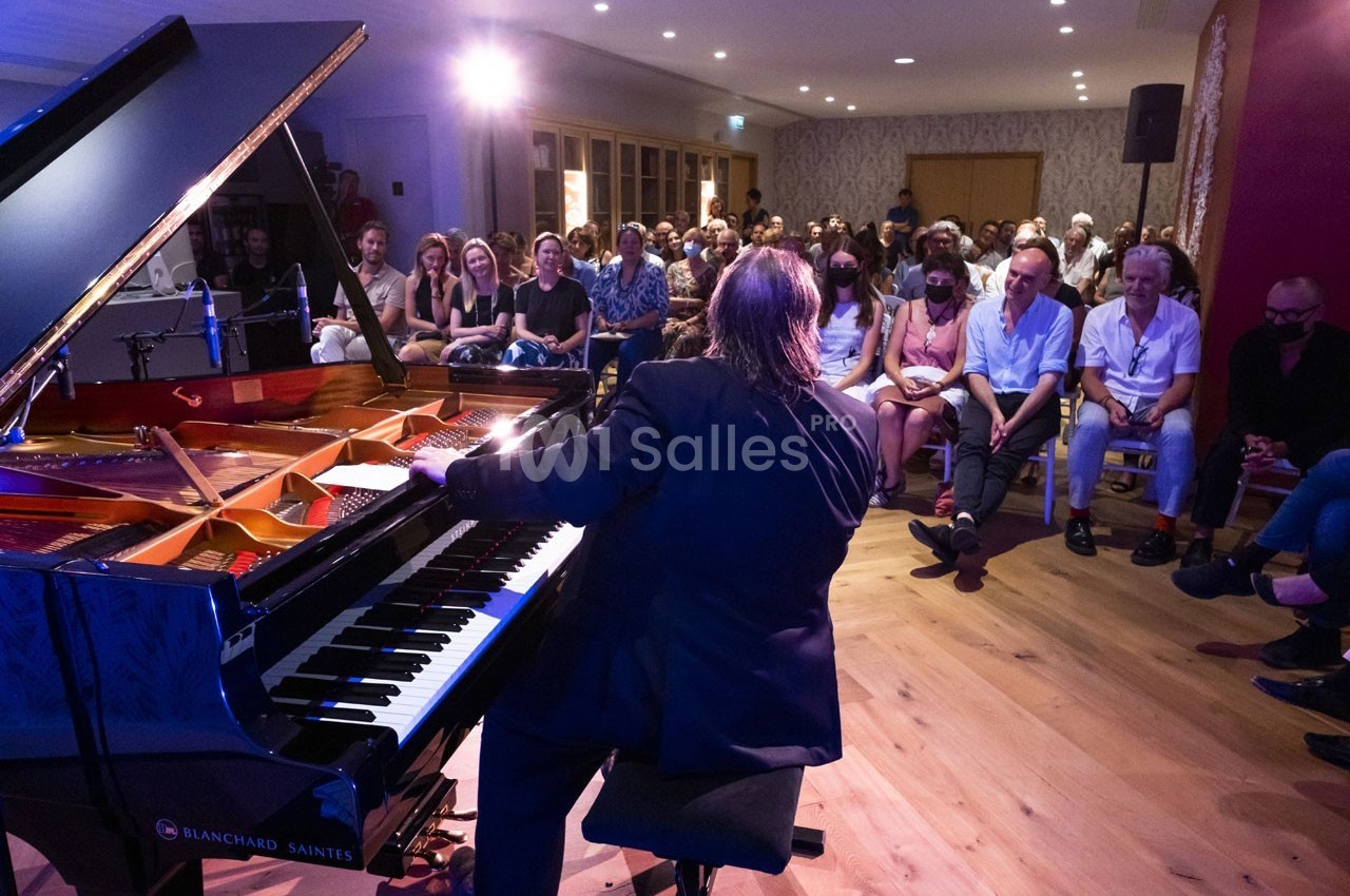 Un pianiste joue devant un public attentif dans une salle lumineuse avec parquet en bois.