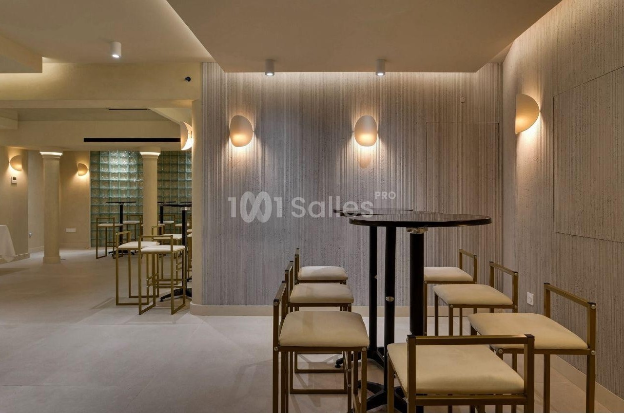 Salle moderne avec des murs texturés, des appliques lumineuses, des tables hautes noires et des chaises à structure dorée.
