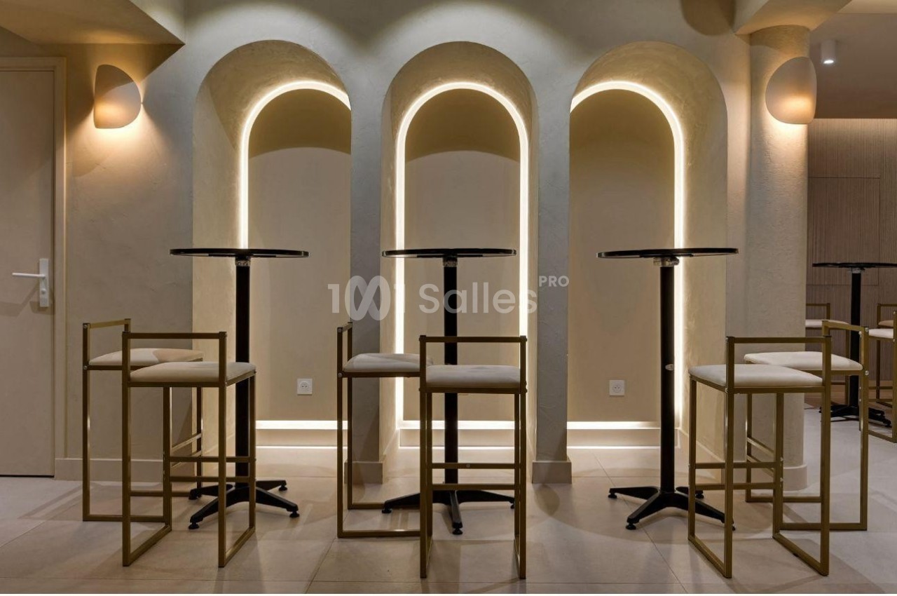 Tables hautes avec tabourets en métal doré devant des arches lumineuses dans un intérieur moderne et épuré.