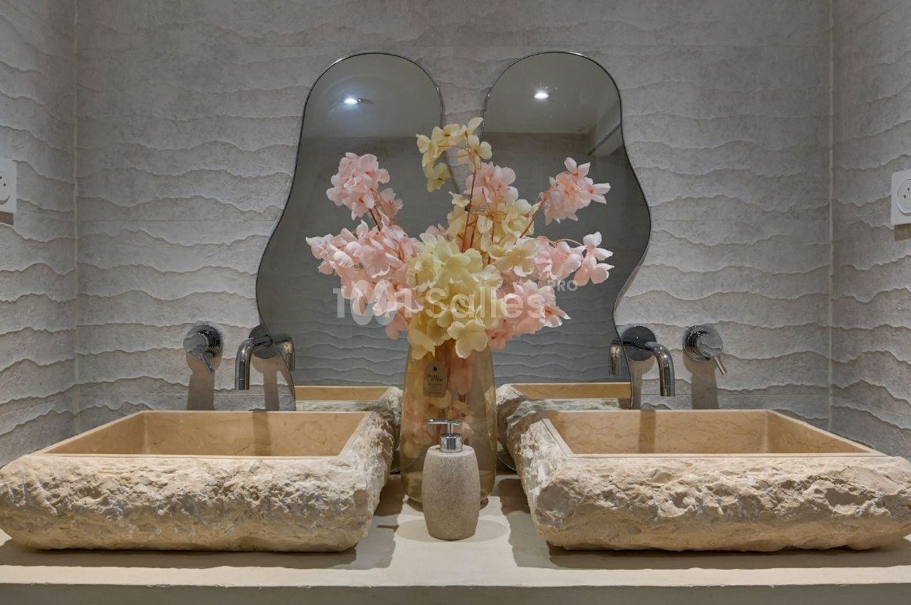 Deux lavabos en pierre avec un vase de fleurs roses et jaunes, placés devant un miroir ondulé.