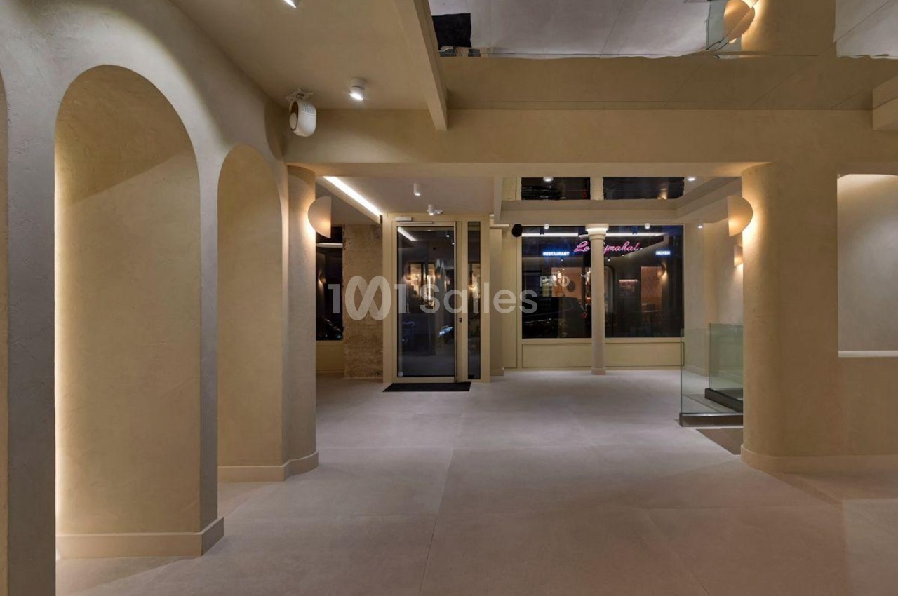 Intérieur d'un espace moderne avec murs beige, arches, éclairage doux et grandes vitrines donnant sur l'extérieur.