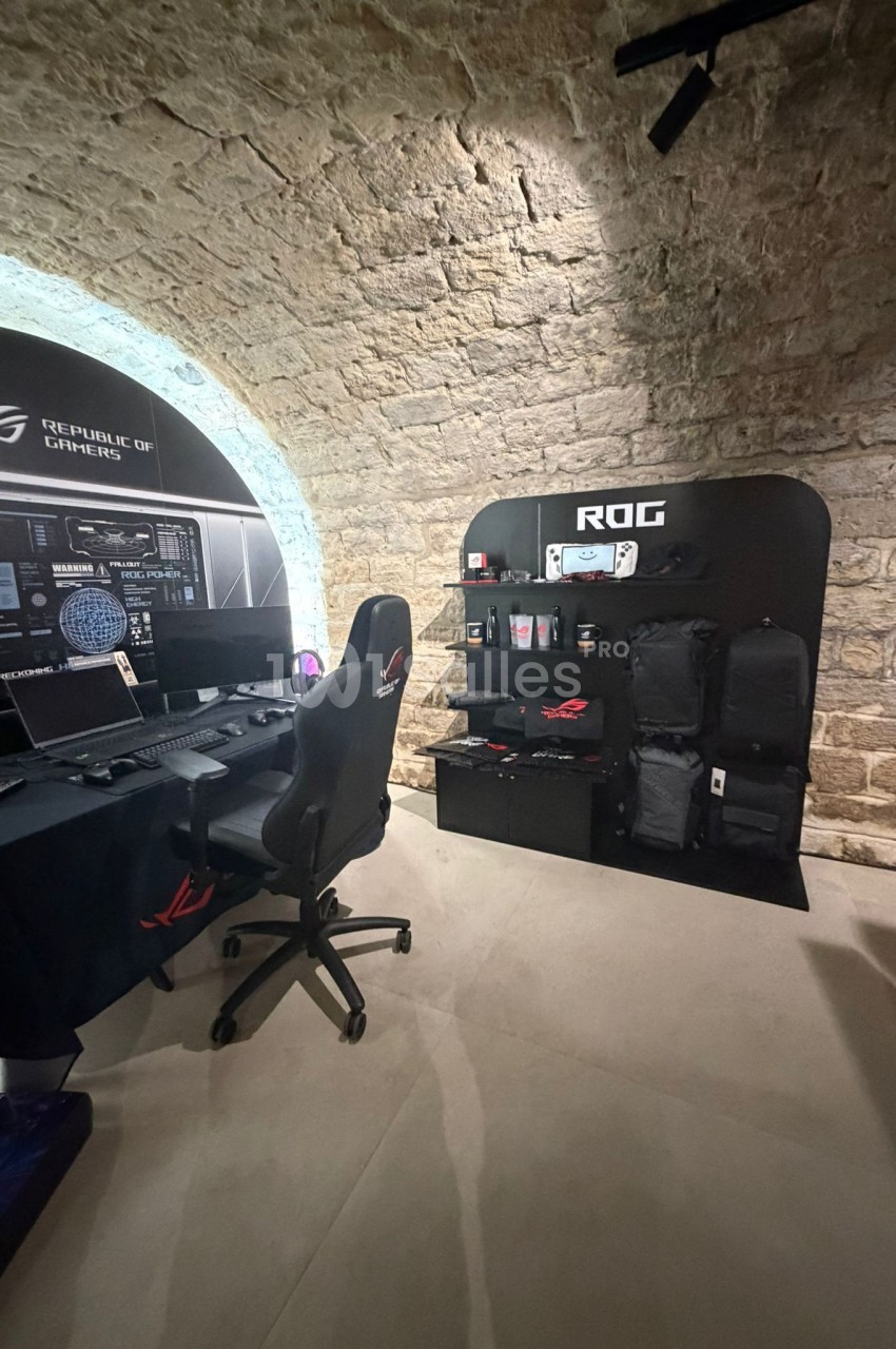Poste de travail gaming avec chaise, écran et accessoires, situé dans une pièce voûtée en pierre avec présentoir de sacs et…