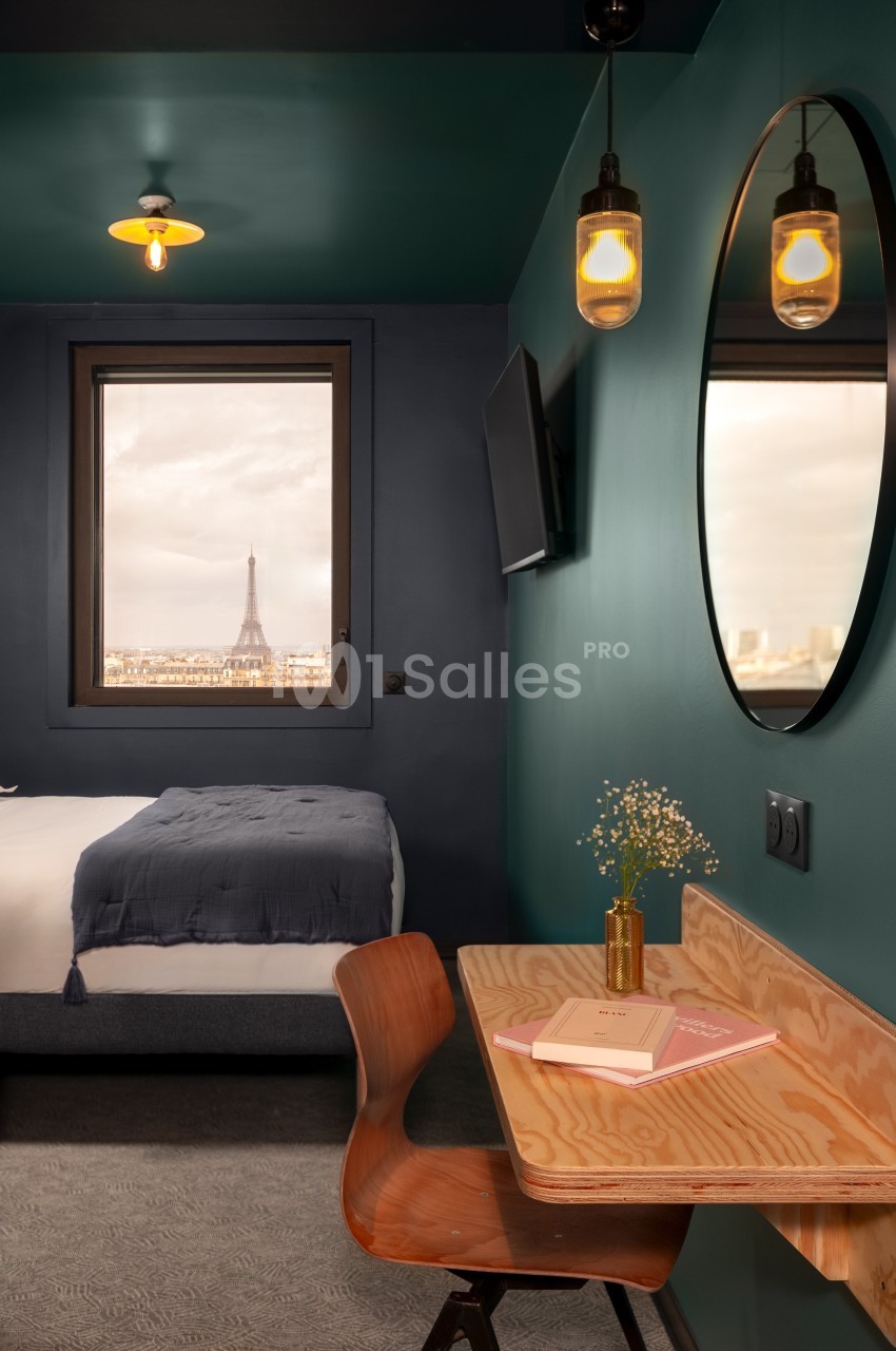 Chambre avec bureau en bois, miroir rond, lit et vue sur la Tour Eiffel par une fenêtre.