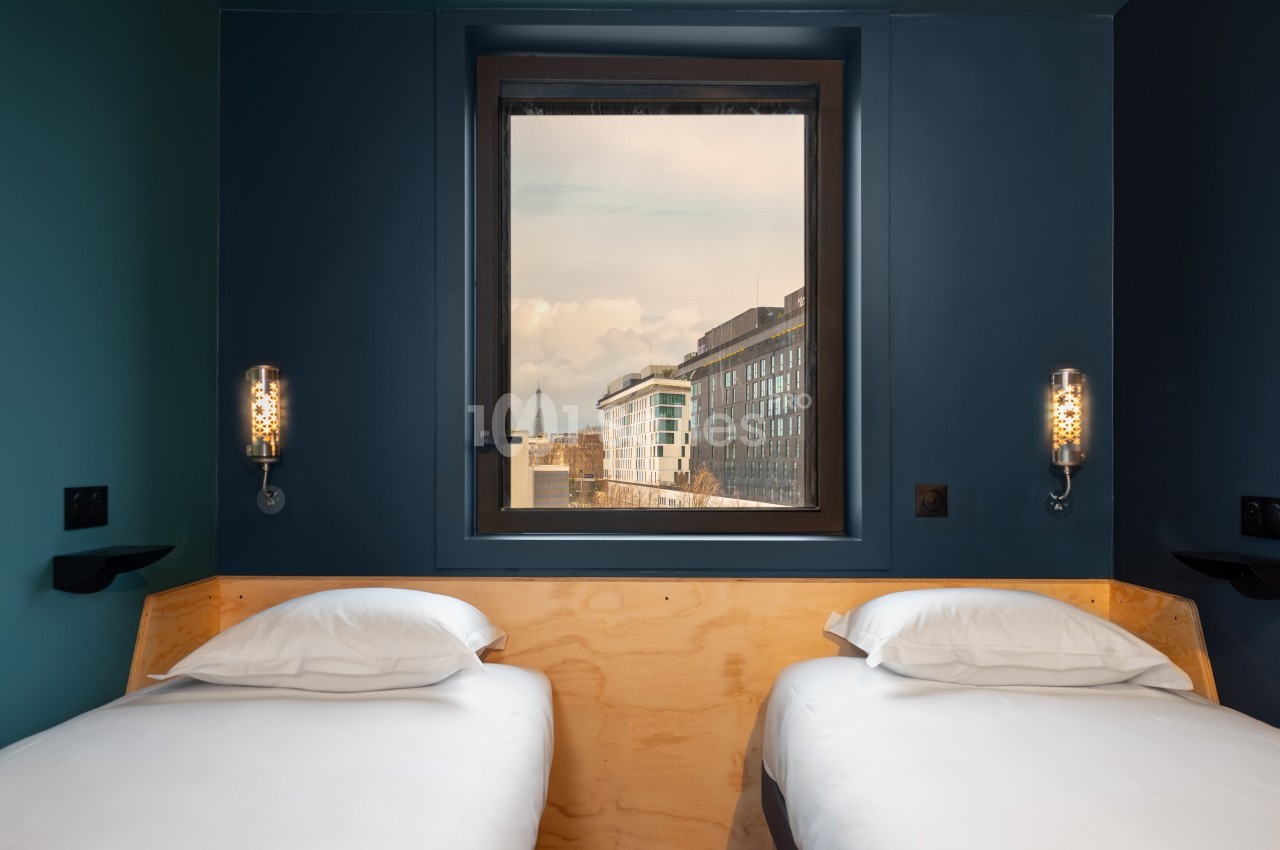 Deux lits simples avec linge blanc dans une chambre aux murs sombres, vue sur des bâtiments modernes par la fenêtre.