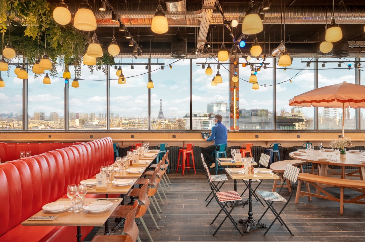 Salle de restaurant lumineuse avec vue sur Paris, tables dressées, banquettes rouges et décoration suspendue.