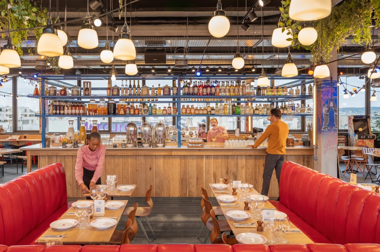 Salle de restaurant moderne avec banquettes rouges, tables dressées, bar en arrière-plan et personnel en préparation.