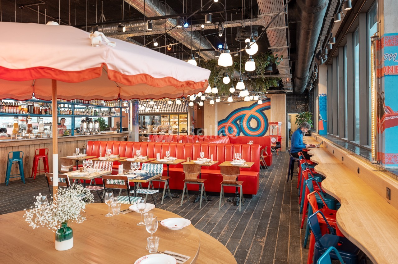 Salle de restaurant lumineuse avec banquettes rouges, tables en bois, chaises colorées et décoration moderne.