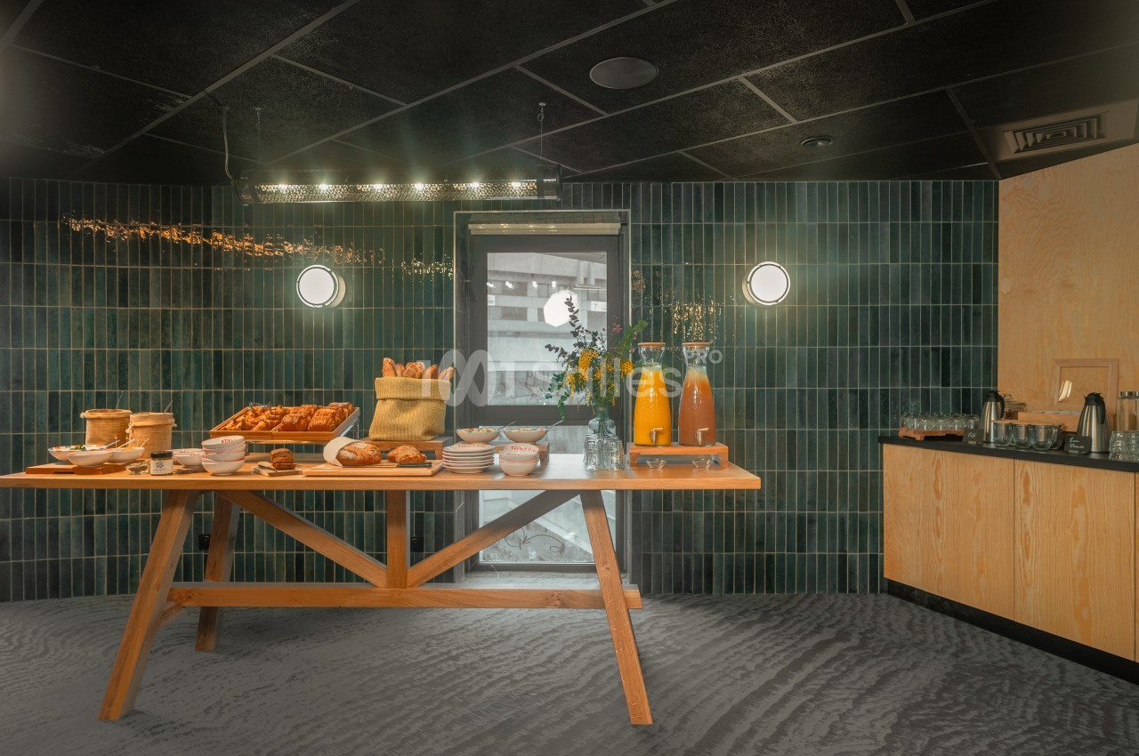 Table en bois dressée avec viennoiseries, pains, jus d'orange et vaisselle, dans une salle aux murs carrelés verts.