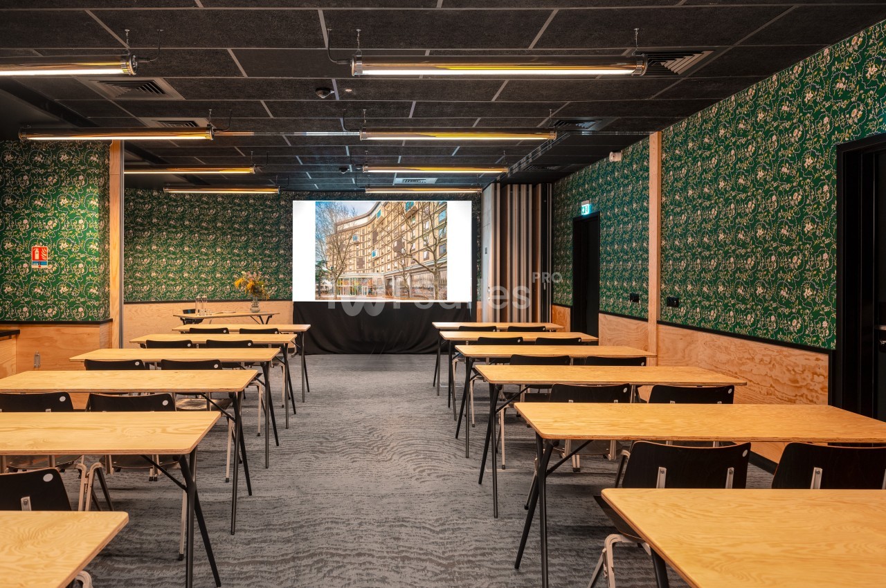 Salle de conférence avec tables en bois clair, chaises noires et écran de projection allumé, décor mural vert à motifs.