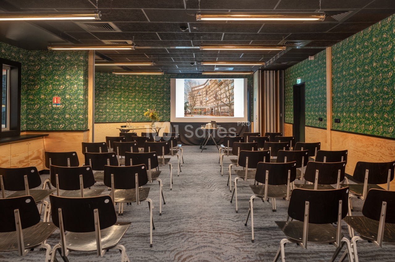 Salle de conférence avec des rangées de chaises noires, un écran de projection allumé et des murs décorés de motifs verts.