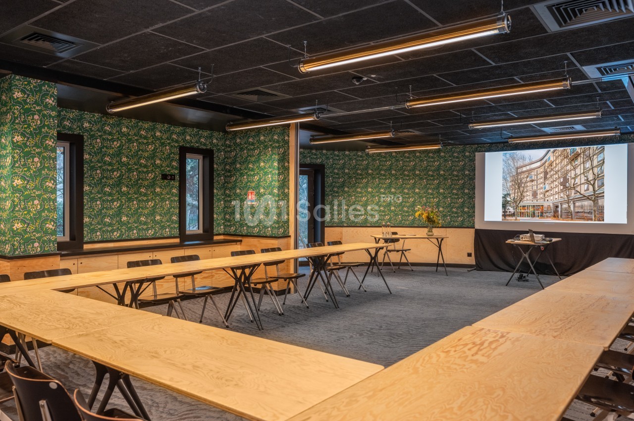 Salle de réunion avec tables en bois disposées en U, écran de projection allumé et murs décorés de papier peint vert.