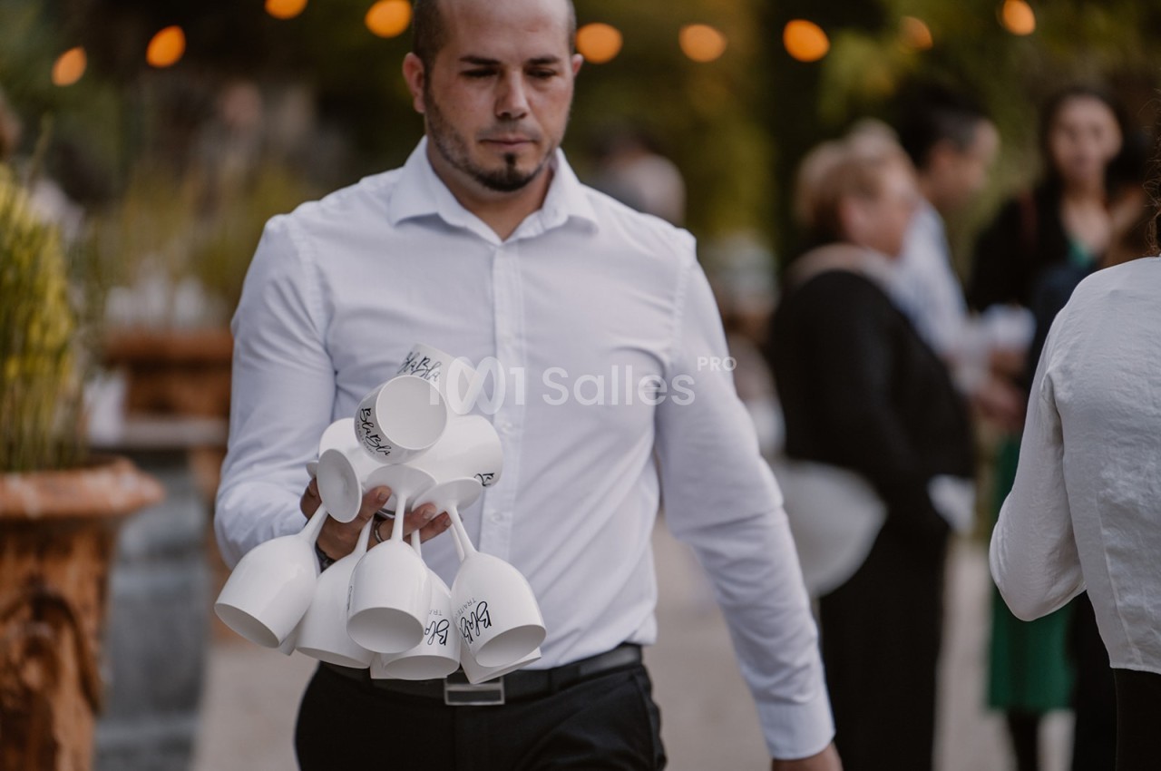 Un homme en chemise blanche porte plusieurs tasses blanches empilées par leurs anses lors d'un événement en extérieur.