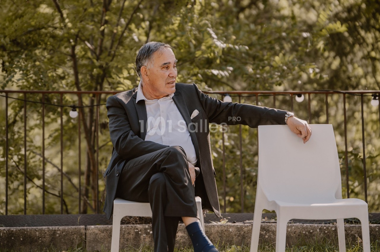 Un homme en costume assis sur une chaise blanche à l'extérieur, près d'une balustrade et entouré de verdure.