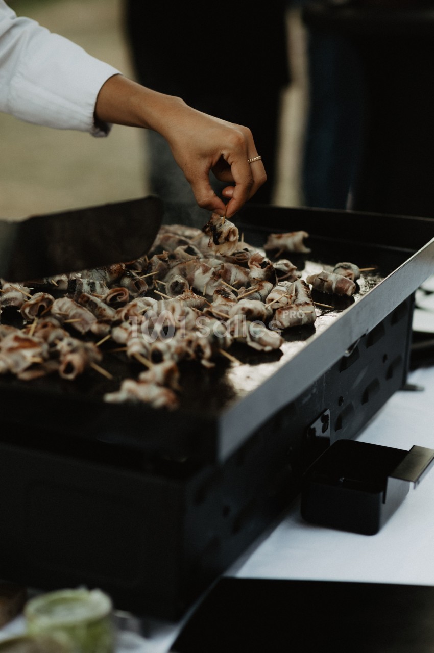 Une main retourne des brochettes de viande grillées sur une plancha noire lors d'une cuisson en extérieur.