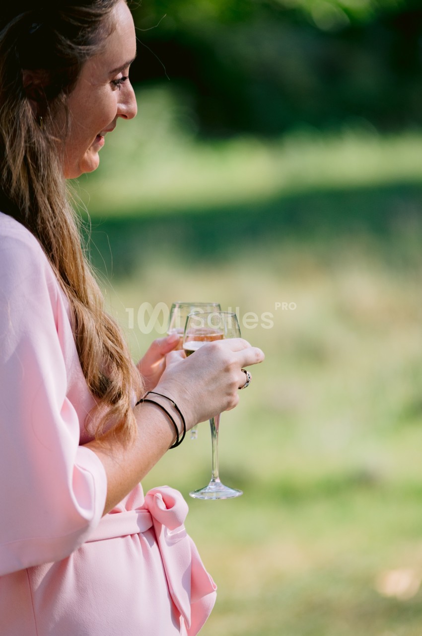 Une femme en robe rose tient un verre de vin blanc dans un jardin ensoleillé.