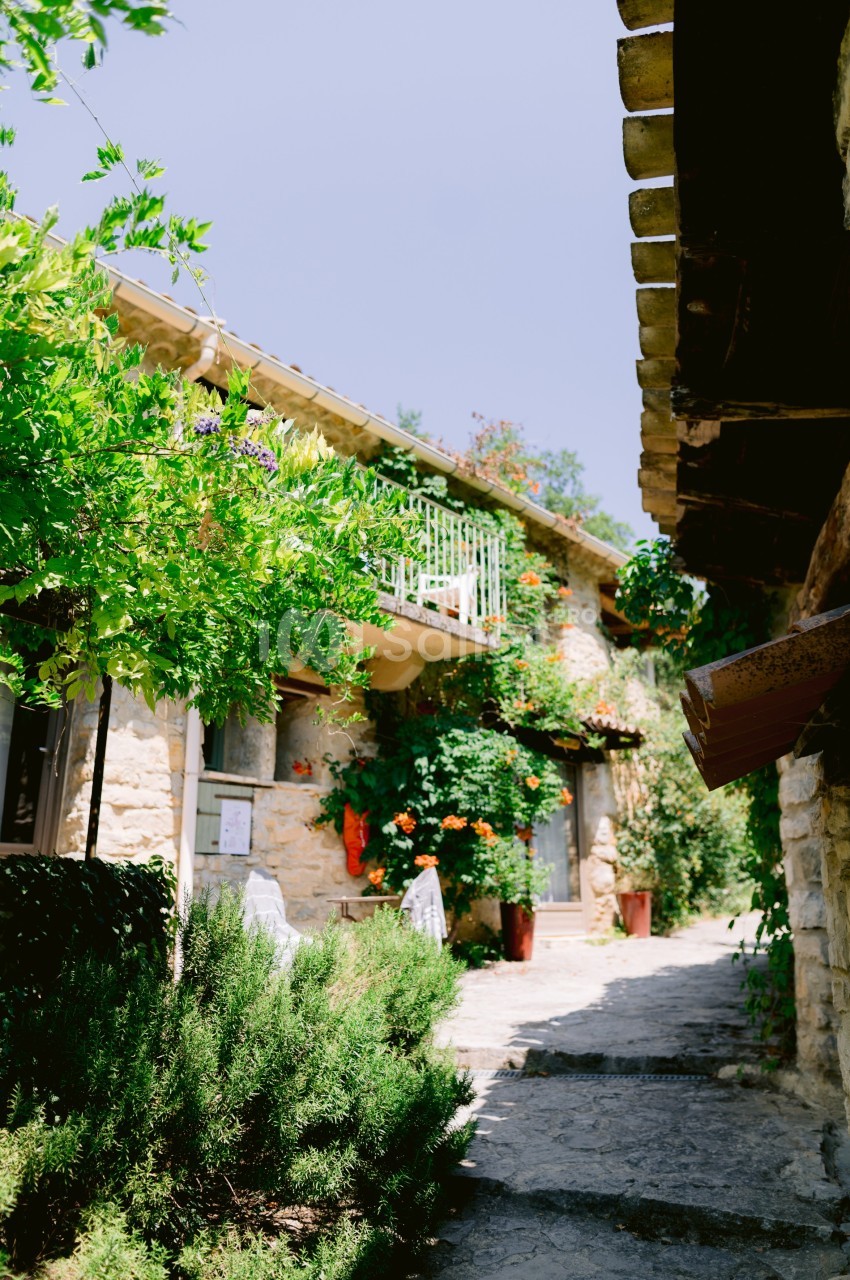 Façade en pierre d'une maison entourée de végétation, avec un balcon fleuri et un chemin en pierre.