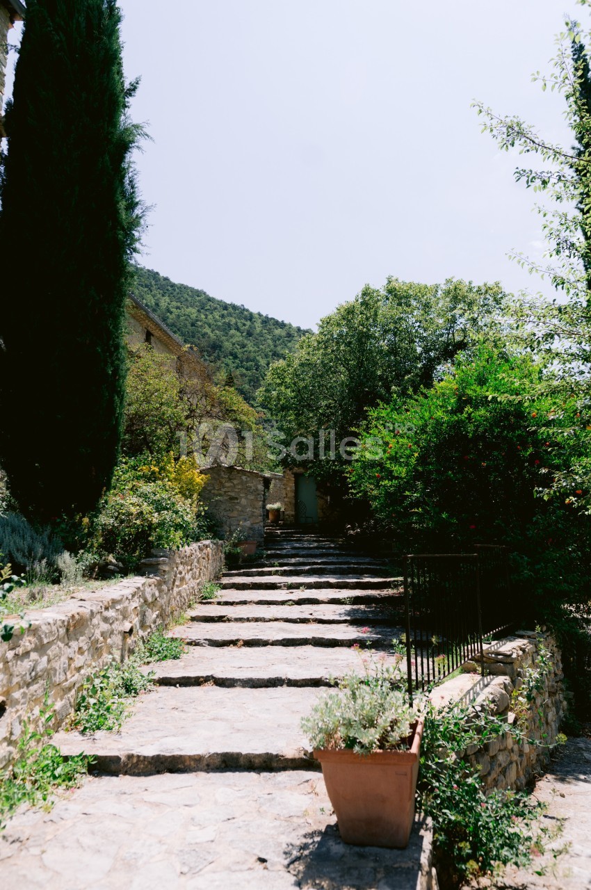 Escalier en pierre bordé de végétation, menant à une maison en pierre dans un cadre naturel ensoleillé.