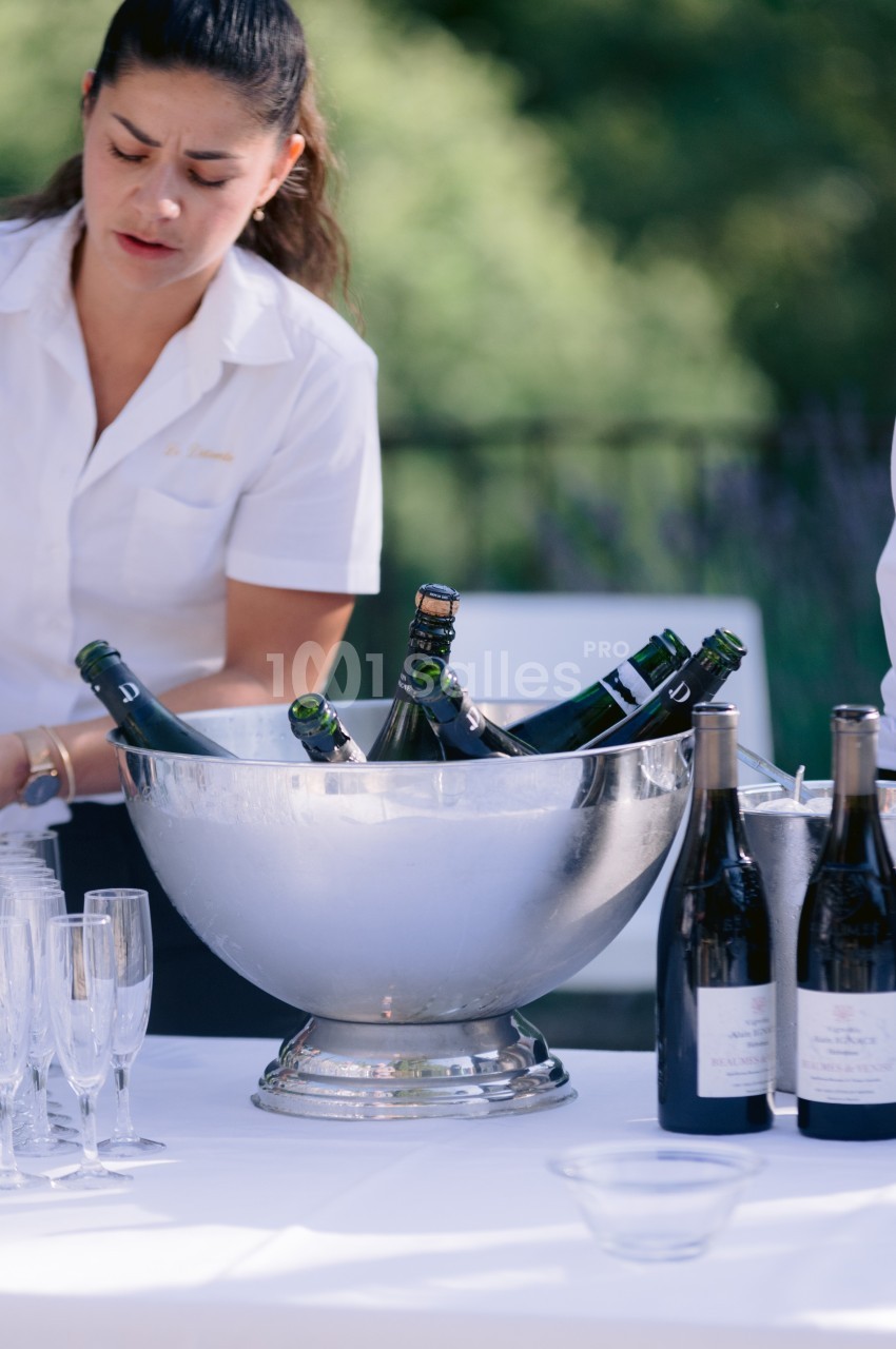 Une femme en chemise blanche prépare une table avec un seau rempli de bouteilles et des verres à champagne.