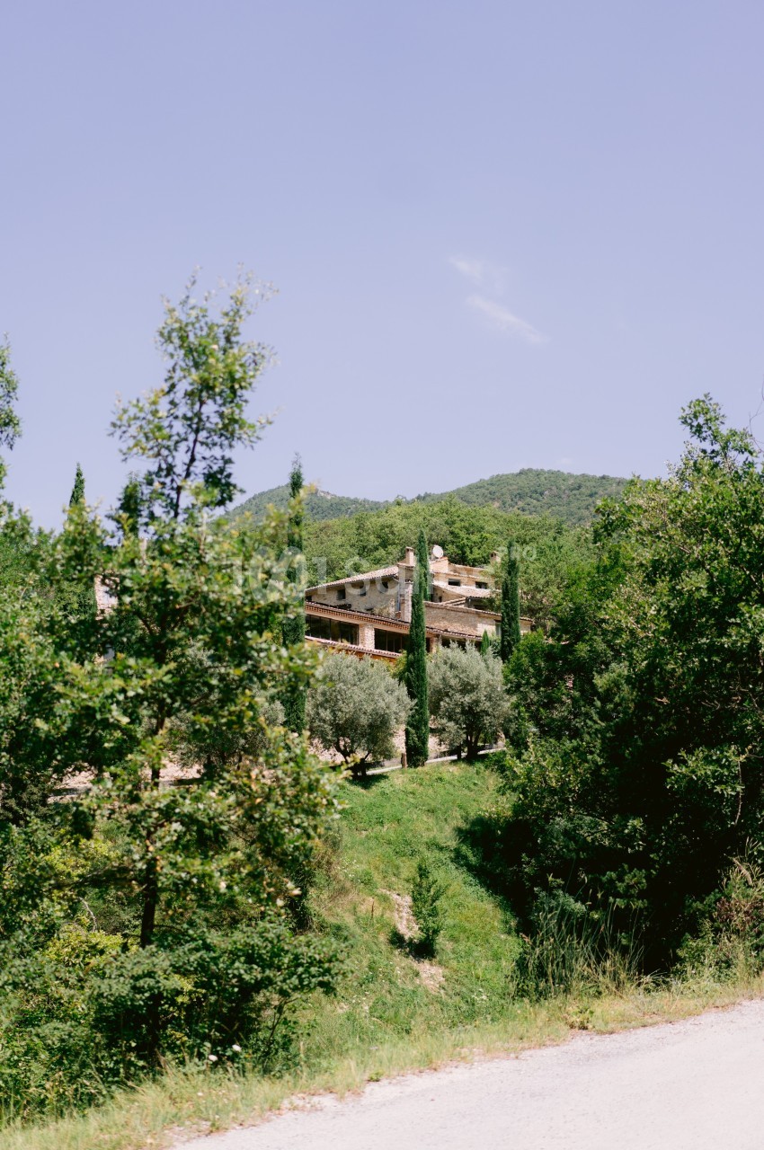 Maison en pierre entourée de cyprès et de végétation, située dans un paysage vallonné sous un ciel dégagé.