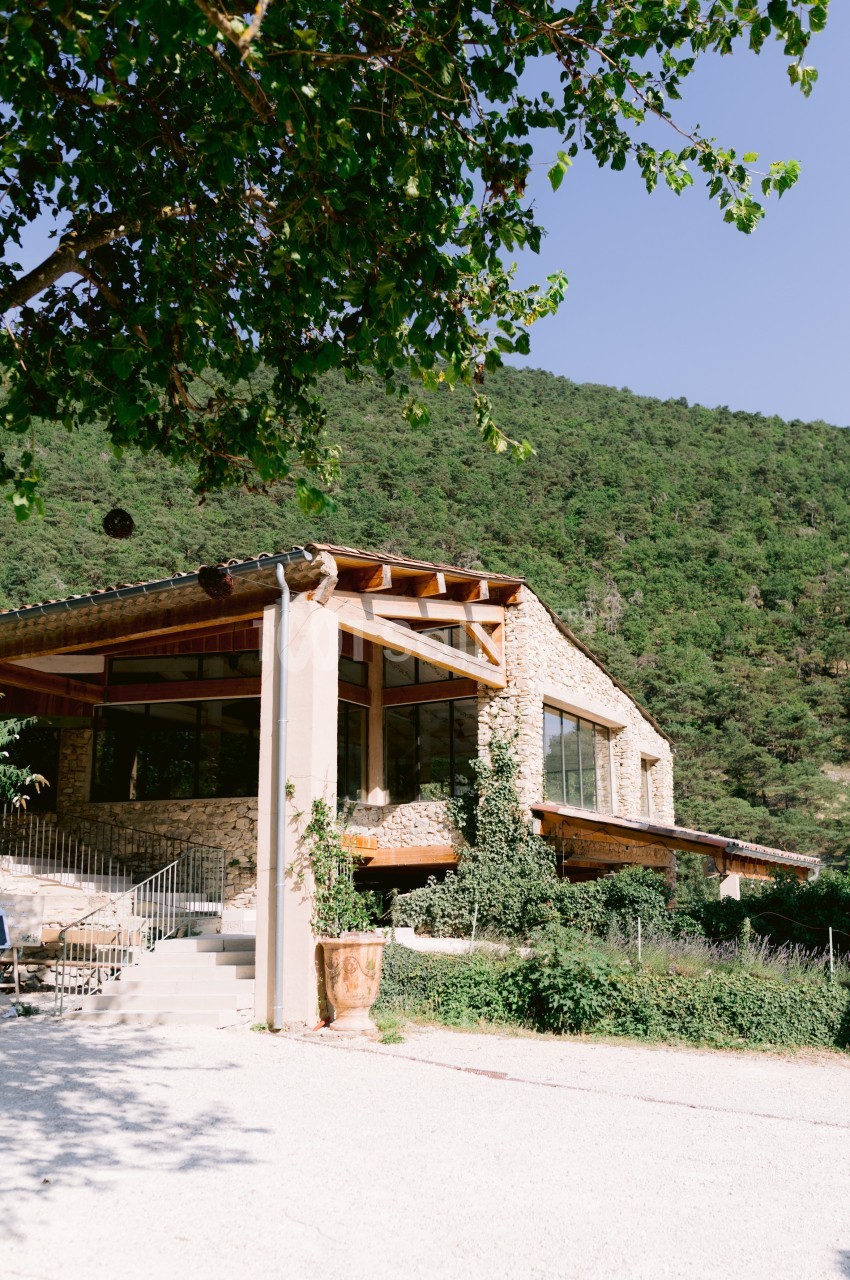 Bâtiment en pierre avec de grandes baies vitrées, entouré de verdure et situé au pied d'une colline boisée.