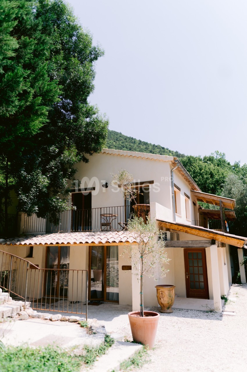 Maison méditerranéenne à deux étages avec balcon, entourée de végétation et éclairée par une lumière naturelle.