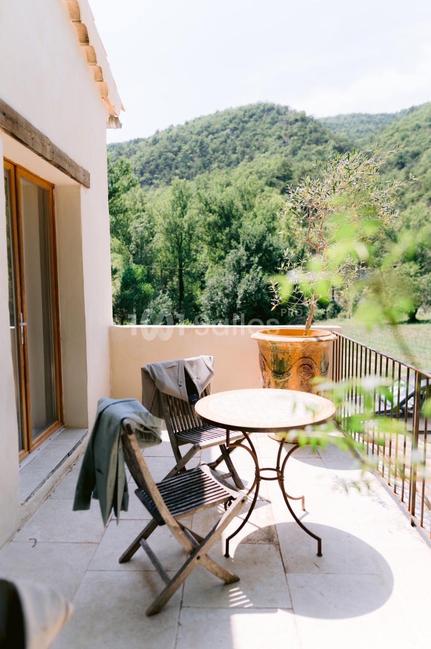 Terrasse ensoleillée avec table ronde, chaises en bois, pot en terre cuite et vue sur des collines verdoyantes.