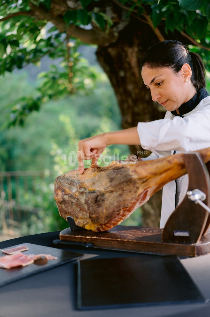 Une femme en tenue de chef découpe un jambon cru sur un support en bois, en extérieur sous un arbre.