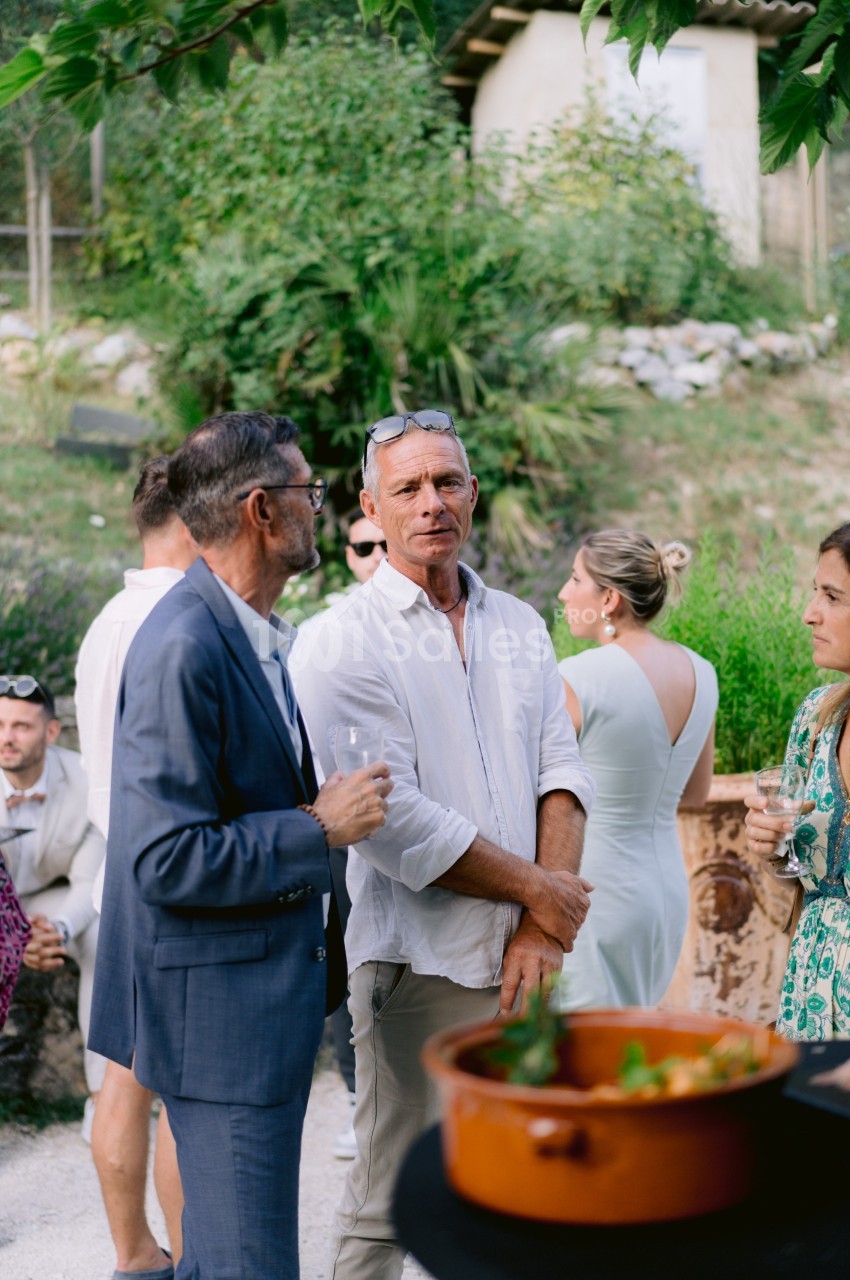 Un groupe de personnes discute dans un jardin, certaines tenant des verres, avec un plat en terre cuite au premier plan.