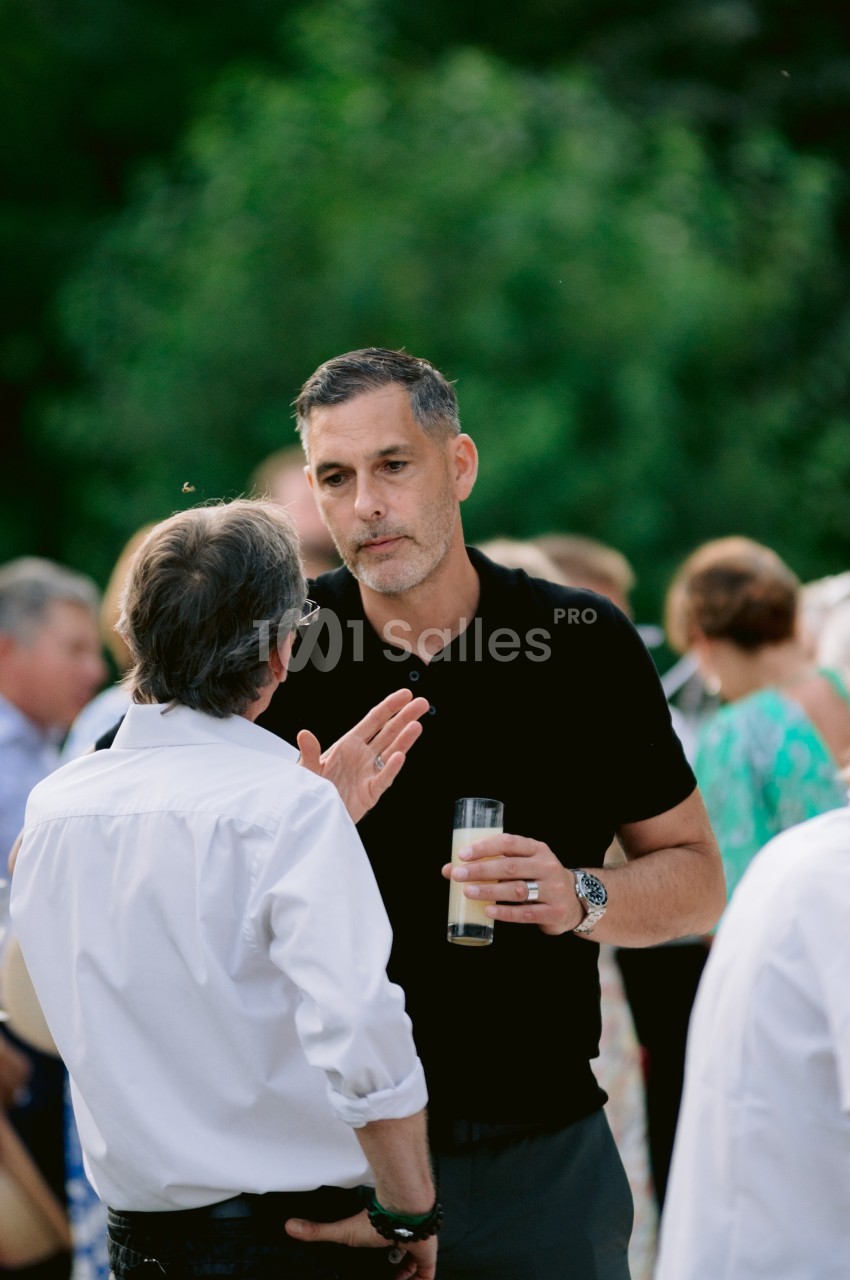 Deux hommes discutent debout lors d'un événement en plein air, l'un tenant un verre à la main.
