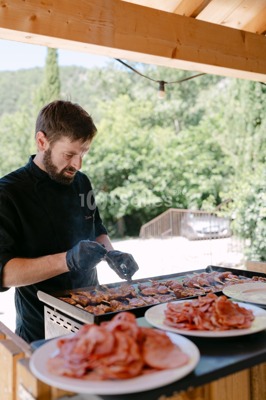 Un cuisinier prépare des brochettes sur un gril en extérieur, avec des assiettes de charcuterie au premier plan.