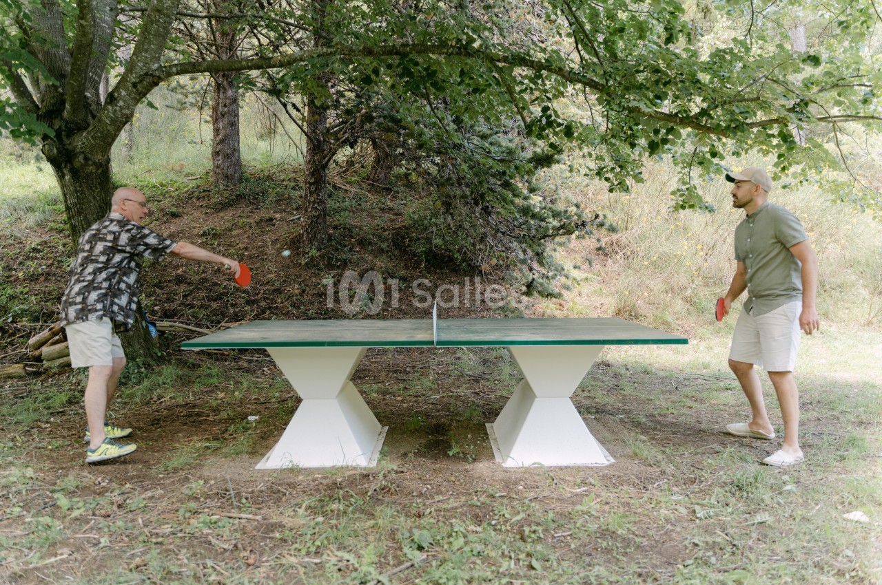 Deux hommes jouent au ping-pong en plein air sur une table située dans un cadre naturel boisé.