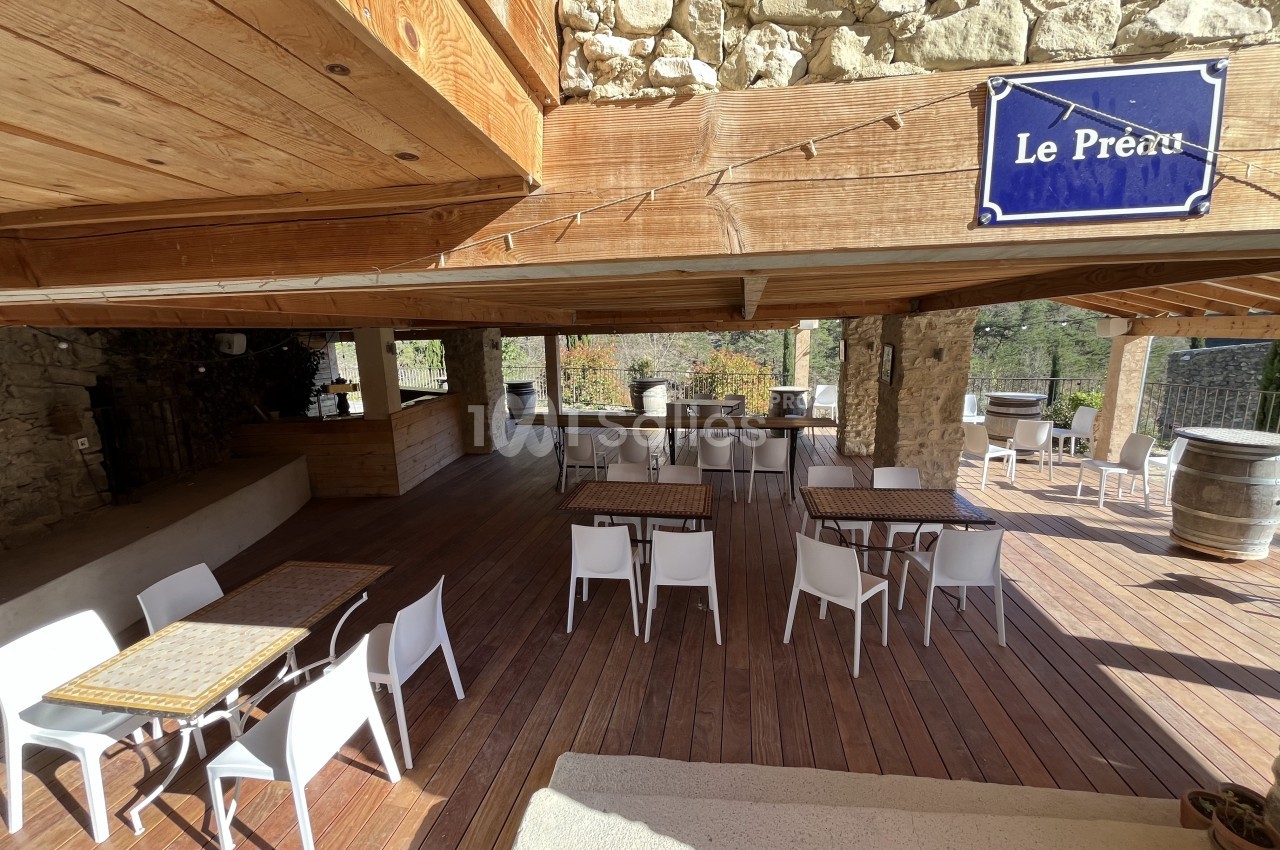 Terrasse en bois avec tables et chaises blanches sous une structure en pierre et bois, éclairée par la lumière naturelle.