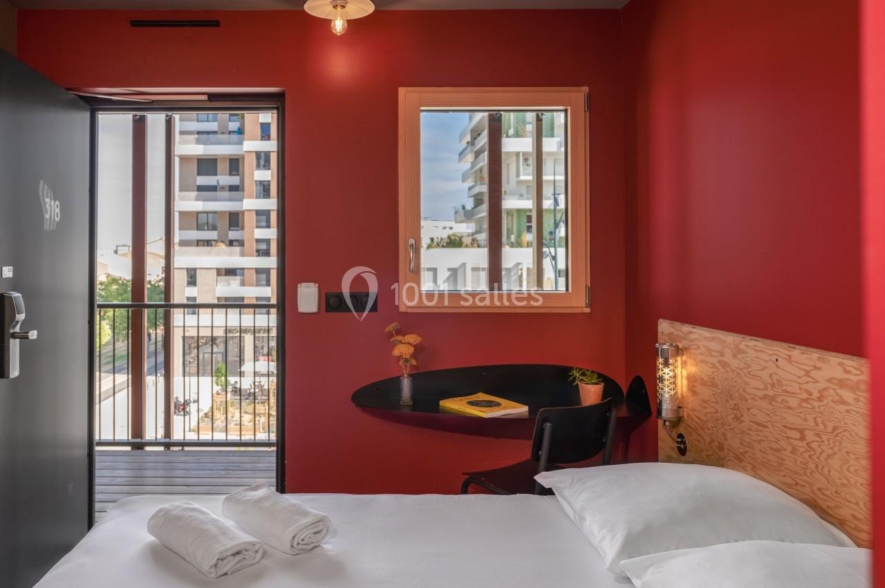 Chambre moderne avec murs rouges, lit double, bureau, fenêtre et balcon donnant sur des immeubles résidentiels.