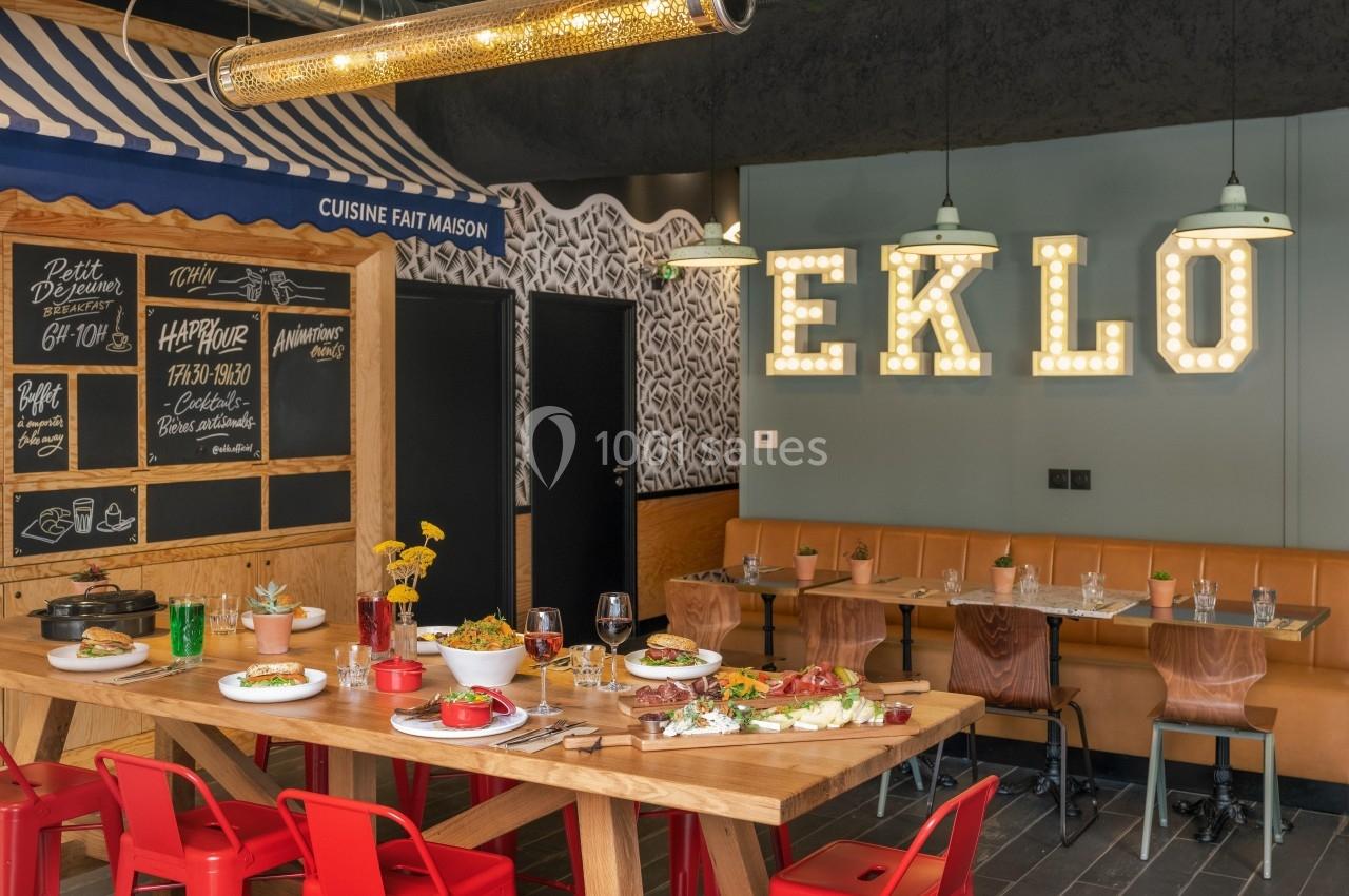 Salle de restaurant moderne avec une grande table dressée, des chaises rouges et une enseigne lumineuse ’EKLO’.