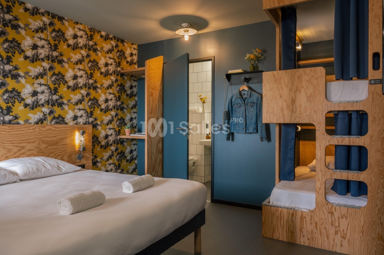 Chambre d'hôtel avec lit double, lits superposés en bois, mur fleuri, et salle de bain visible à travers une porte ouverte.