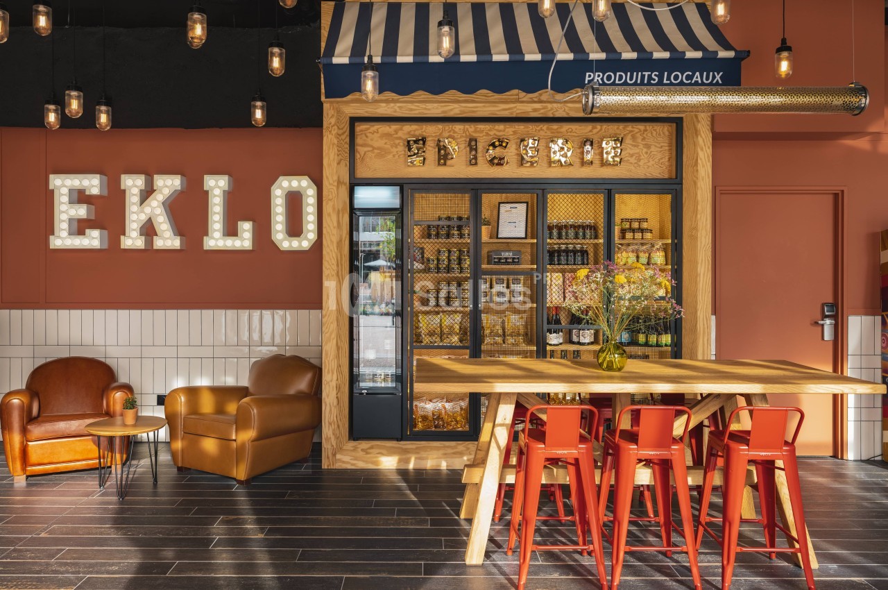 Coin boutique avec étagères de produits locaux, table en bois, chaises rouges et fauteuils dans un décor chaleureux.