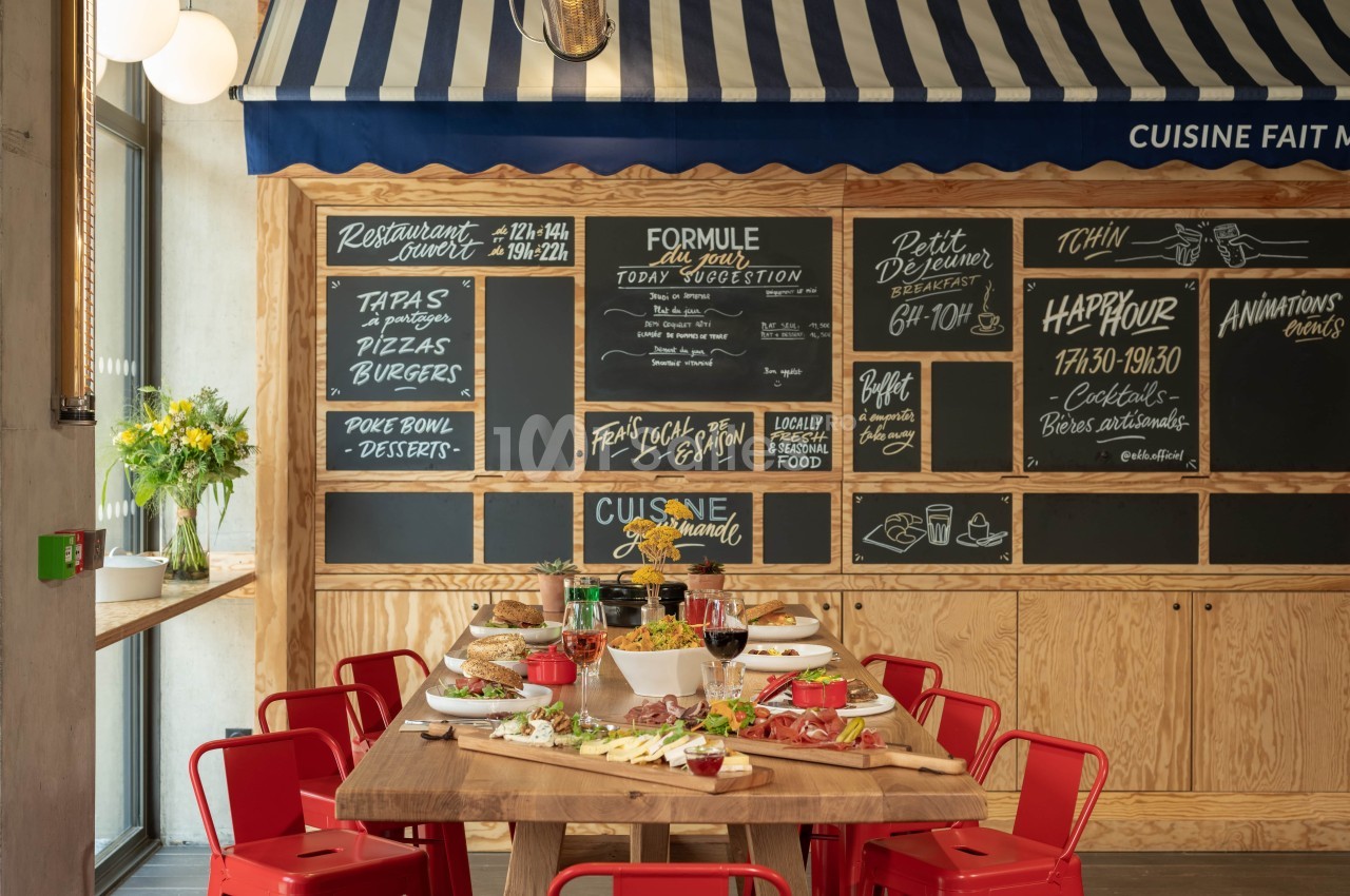 Salle de restaurant avec table en bois dressée pour un repas, ardoises murales affichant menus et offres, chaises rouges.