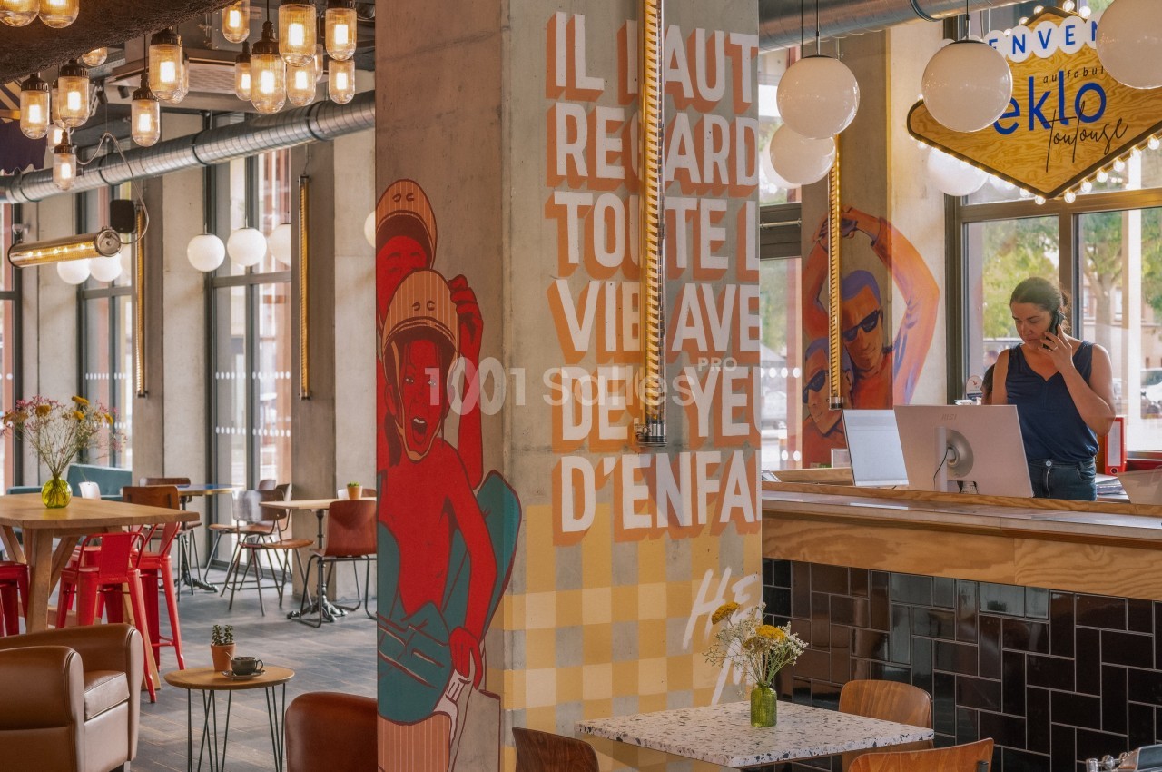 Intérieur d'un café moderne avec une fresque colorée, un comptoir en bois et une personne au téléphone derrière le comptoir.
