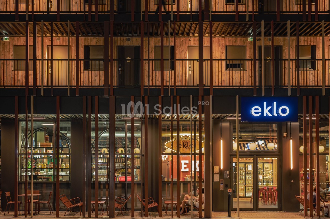 Façade d'un bâtiment moderne avec enseigne lumineuse ’eklo’, terrasse aménagée et intérieur éclairé visible.