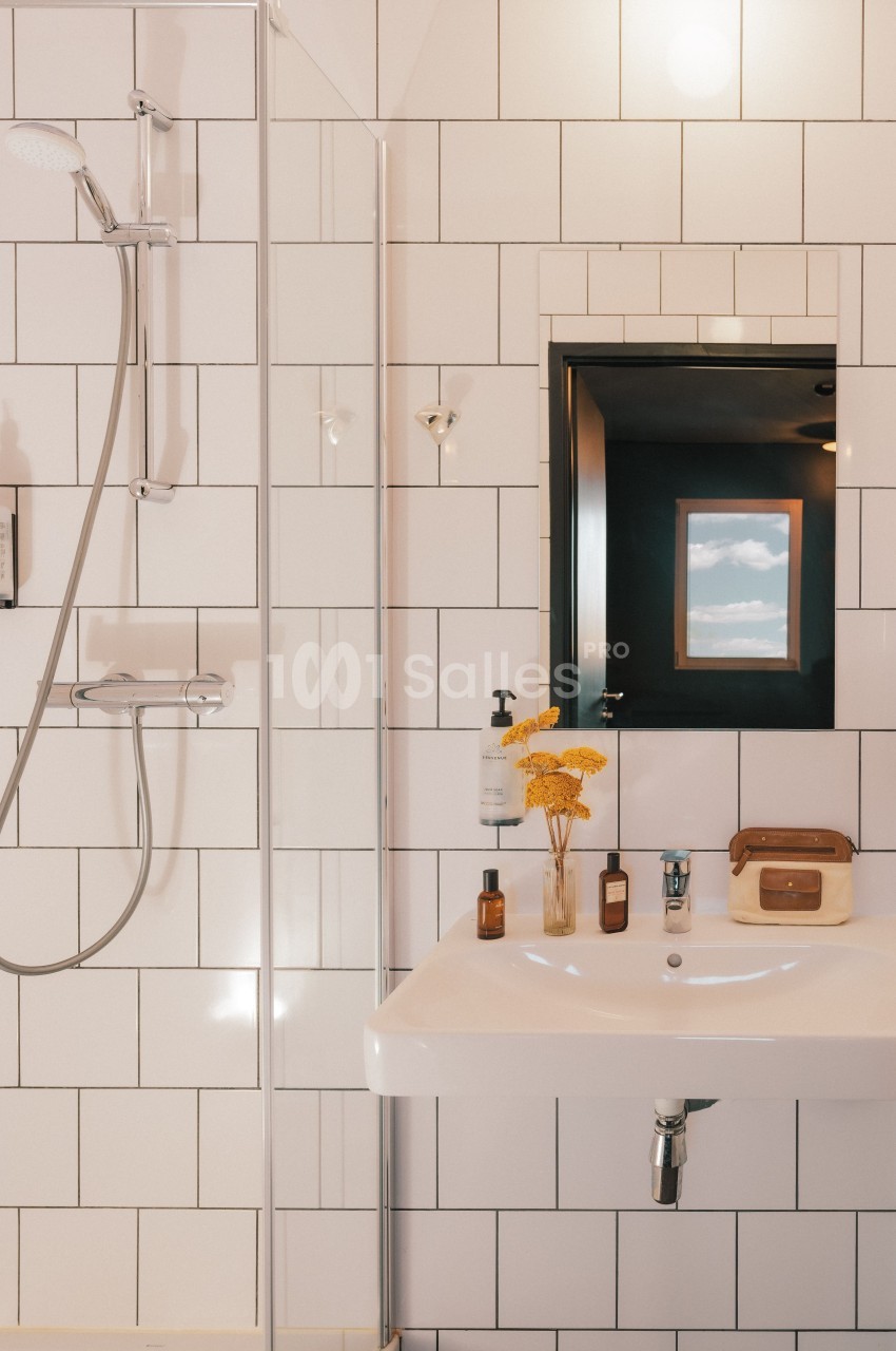 Salle de bain avec douche, lavabo, miroir, flacons et fleurs jaunes, carrelage blanc et fenêtre en arrière-plan.
