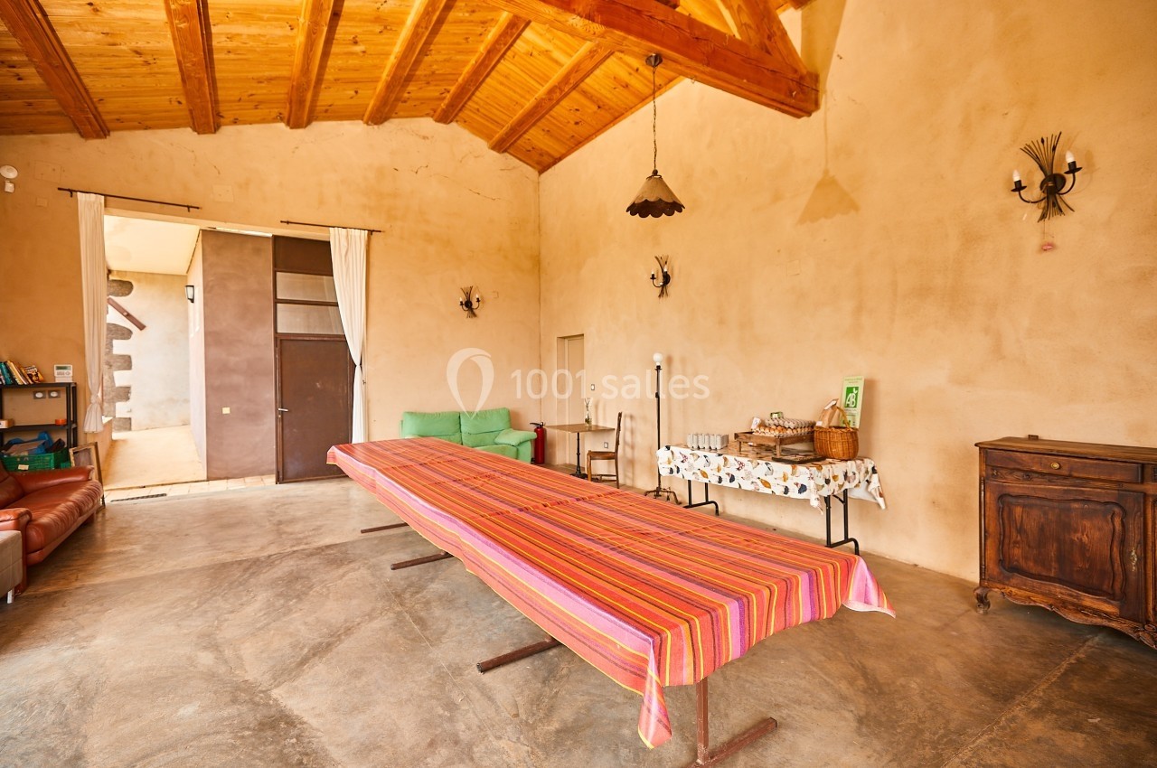 Salle lumineuse avec plafond en bois, grande table avec nappe colorée, buffet avec objets et canapé en fond.