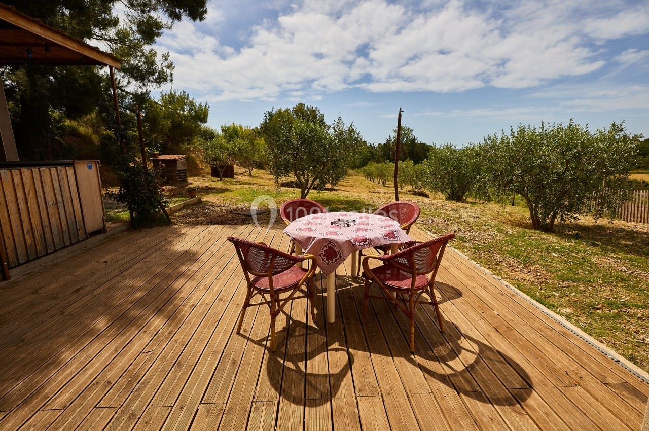 Table ronde avec nappe fleurie et chaises en osier sur une terrasse en bois, entourée d'arbres et de verdure.