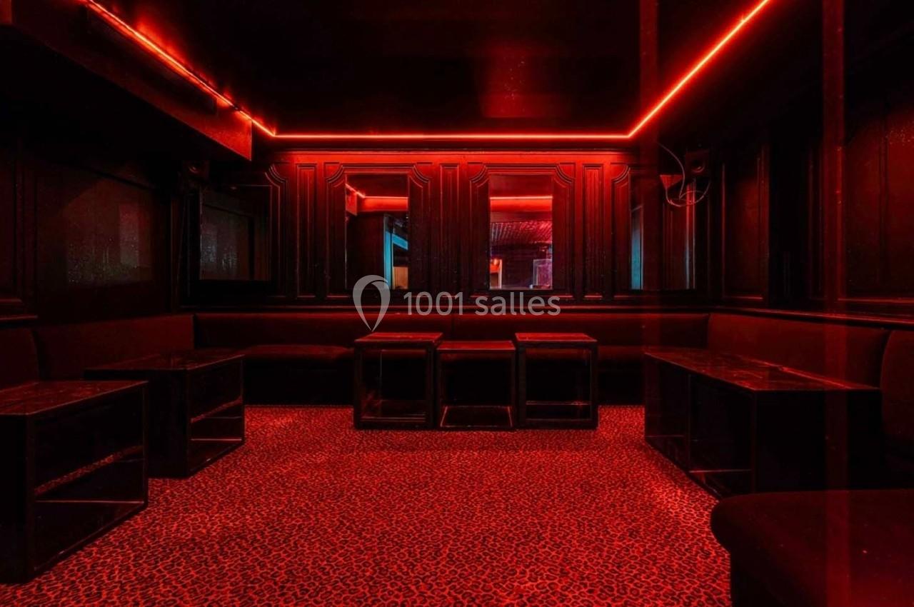 Salle sombre avec éclairage rouge, tables basses noires, banquettes et sol à motif léopard.