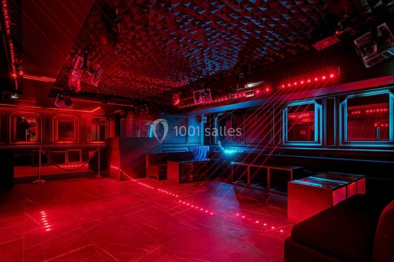 Salle de club avec éclairage rouge et bleu, murs texturés, banquettes et tables basses modernes.