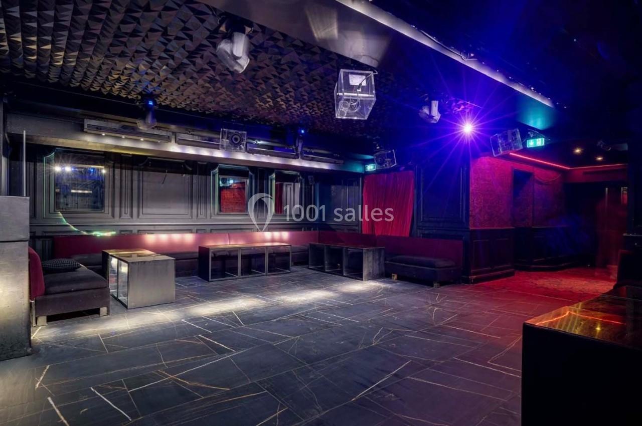 Salle de club vide avec éclairage tamisé, banquettes noires, murs sombres et plafond texturé.