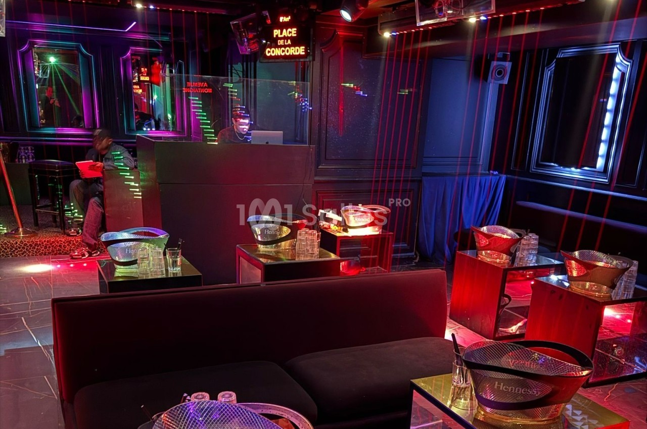 Salle de club avec banquettes, tables basses, seaux à glace et éclairage tamisé aux tons rouges et violets.