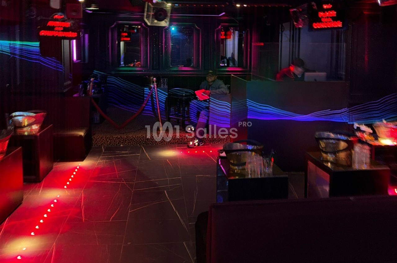 Intérieur d'une discothèque sombre avec éclairages rouges et bleus, un DJ au fond et quelques tables disposées.
