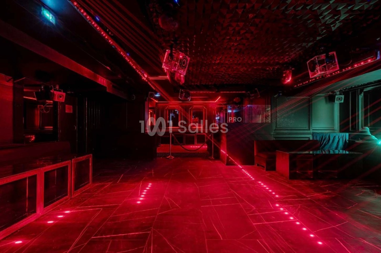 Salle de club vide avec éclairage rouge, murs sombres, sol texturé et équipements audio visibles.